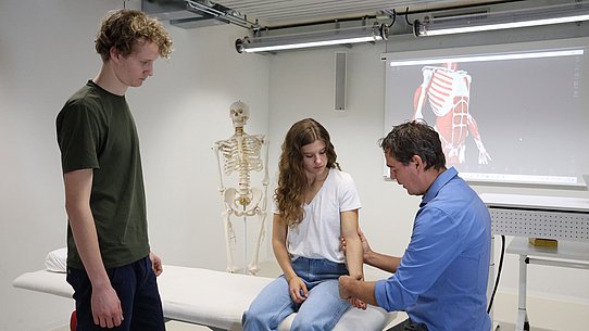 Zwei Studierende und Dozent in Klassenraum. 